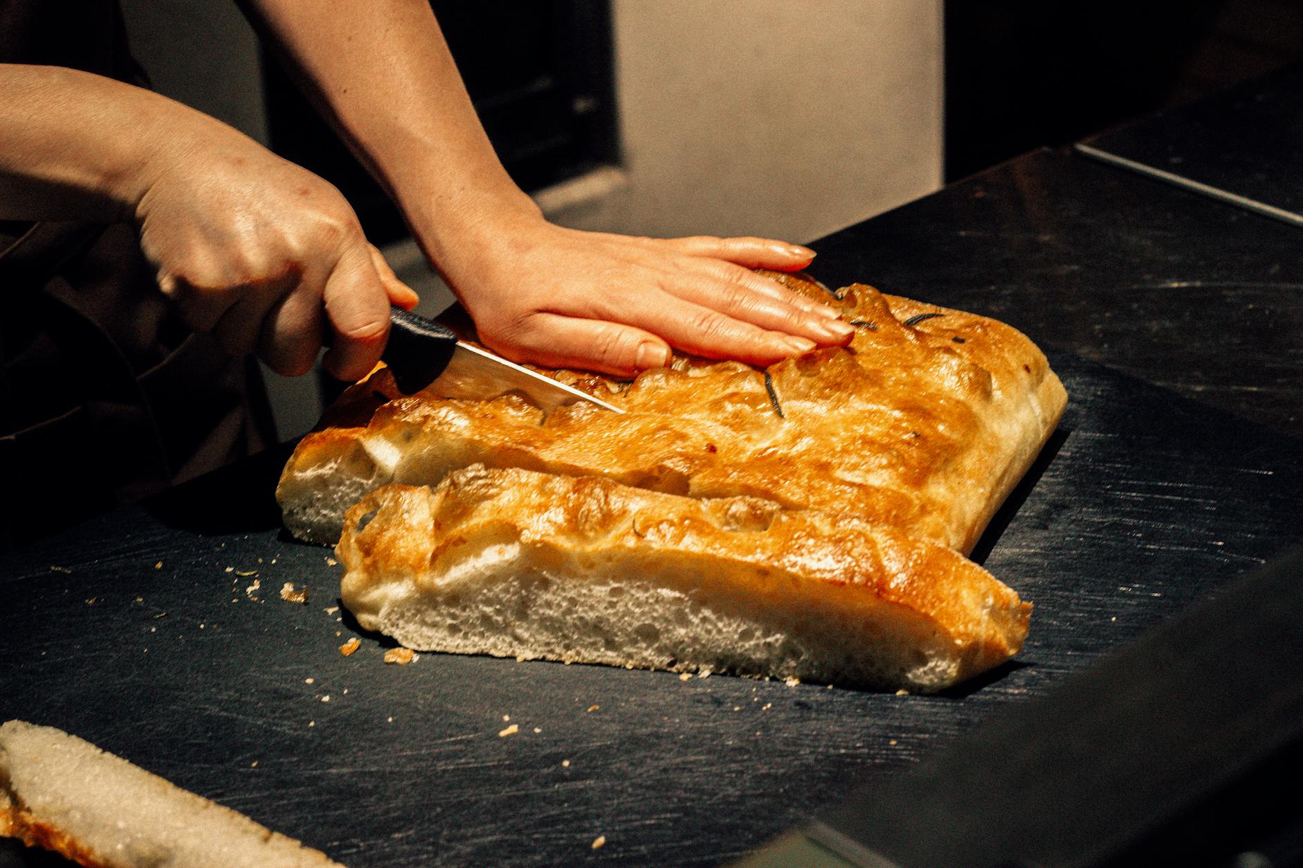 Ktoś kroi świeżą prostokątną focaccię nożem na ciemnym blacie